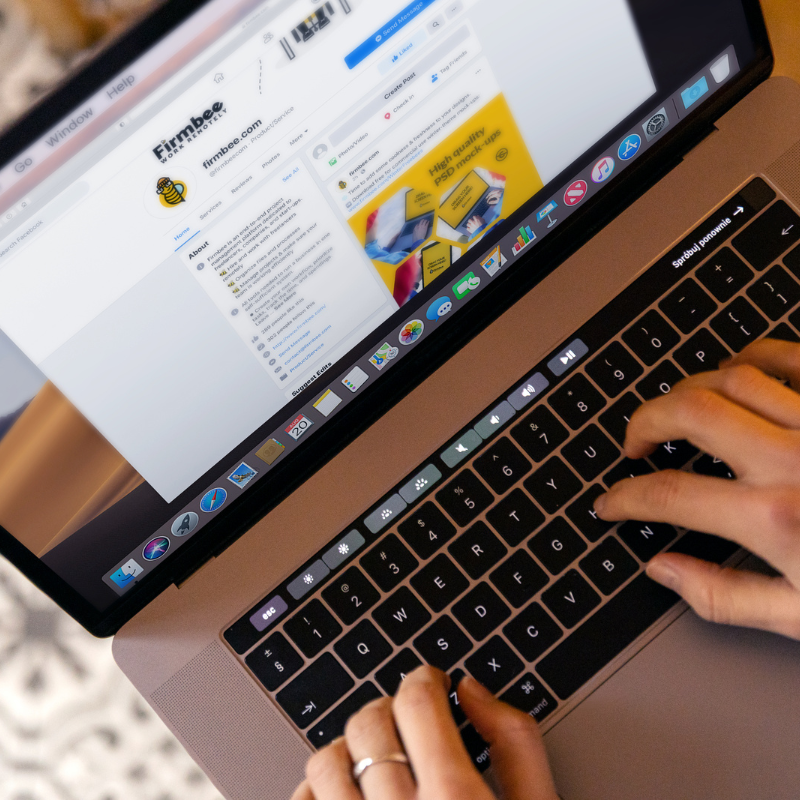 Vue plongeante sur des mains tapant sur le clavier d'un ordinateur portable rose doré. L'écran affiche la page Facebook professionnelle d'une entreprise, avec son logo, sa présentation, ses publications et un visuel promotionnel jaune, tandis que la barre d'applications est visible en bas de l'écran.