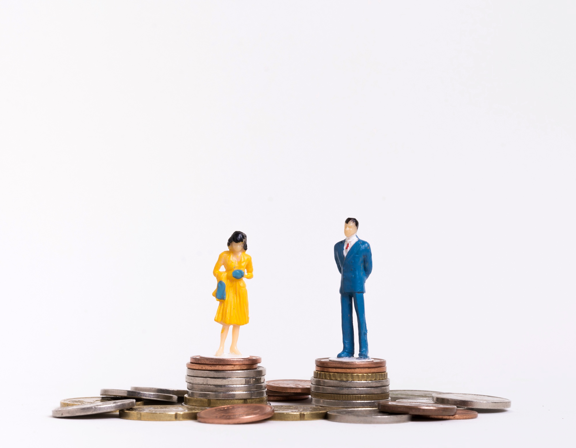 Deux figurines miniatures sur fond blanc : à gauche, une femme en robe jaune tenant un sac et des gants bleus, à droite, un homme en costume bleu, les mains dans le dos. Chacun se tient sur sa propre pile de pièces de monnaie, entourée d'autres pièces éparpillées, évoquant la comparaison des rémunérations entre femmes et hommes.