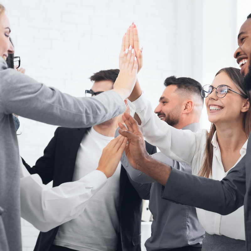Groupe de six collègues en tenue professionnelle, réunis en cercle dans un bureau lumineux aux murs blancs. Ils se tapent dans les mains avec enthousiasme, plusieurs d'entre eux riant aux éclats, dans une ambiance de célébration et de cohésion d'équipe.
