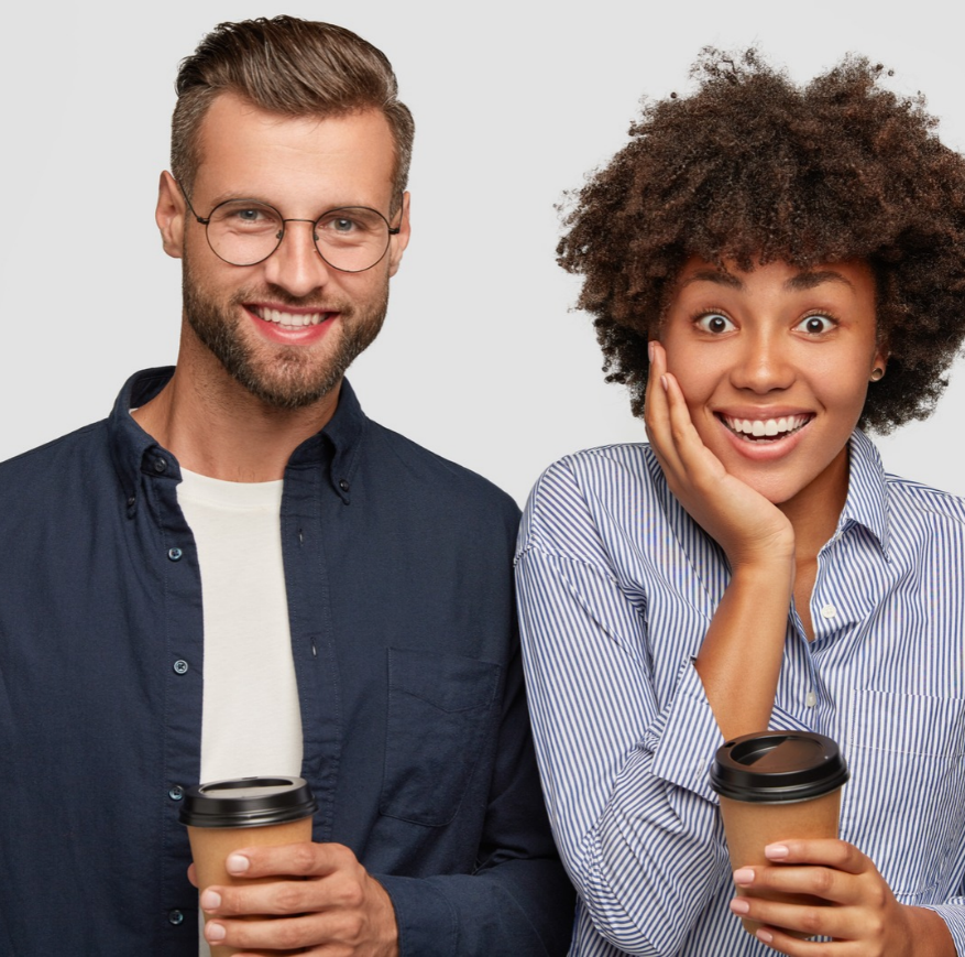 Sur un fond gris clair uni, un homme barbu à lunettes rondes, en chemise bleu marine sur un tee-shirt blanc, sourit en tenant un gobelet de café à emporter. À côté de lui, une femme aux cheveux bouclés, en chemise rayée bleue et blanche, pose la main sur sa joue, les yeux grands ouverts et un large sourire aux lèvres, un café à la main elle aussi.